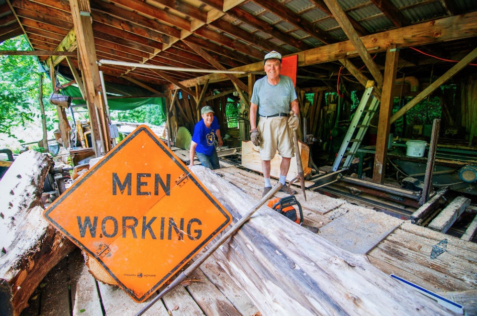 Two men smiling inside the open-sided Walker's Sawmill shed, with rough-sawn boards in the foreground